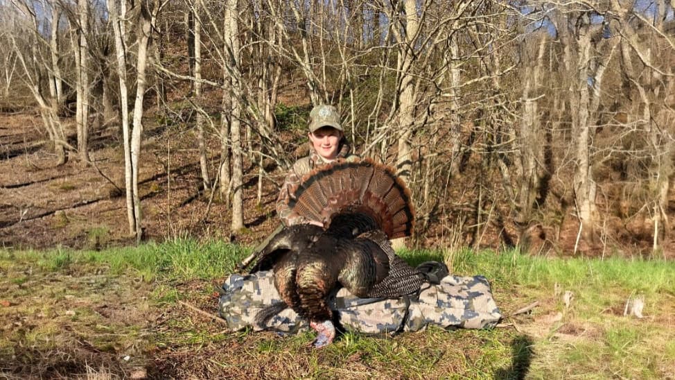 Young person wearing camo hat and clothing holds up a large turkey, its tail fanned out. The bird is on a camouflaged bag...