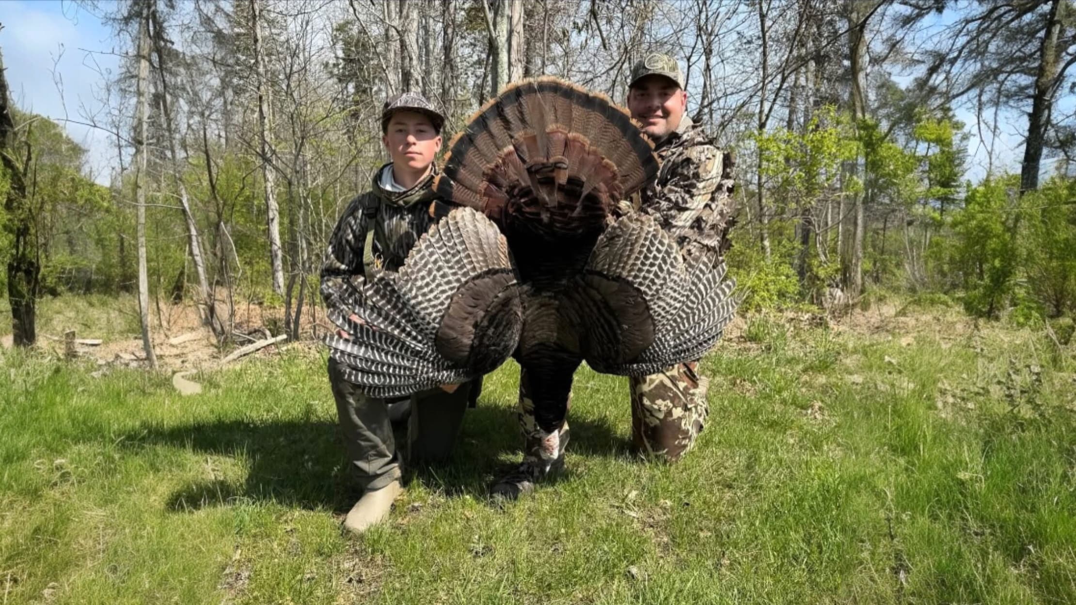 Two men in camo holding a large turkey in a forest. The turkey's tail feathers are fanned out. Green grass and trees are i...