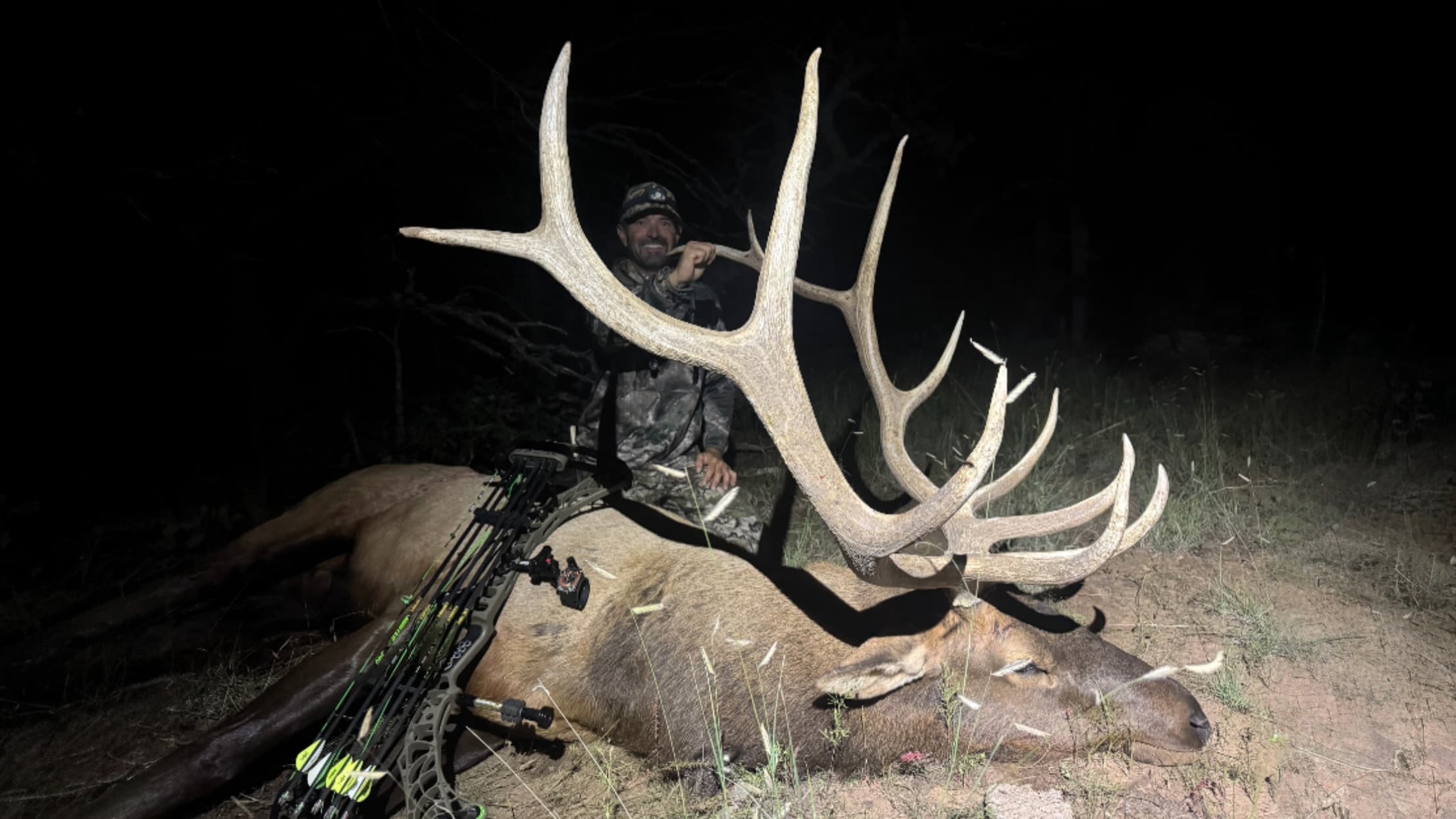 Hunter kneeling next to a harvested elk with large antlers in the dark. A bow and arrows lie nearby. Hunter smiles at the ...