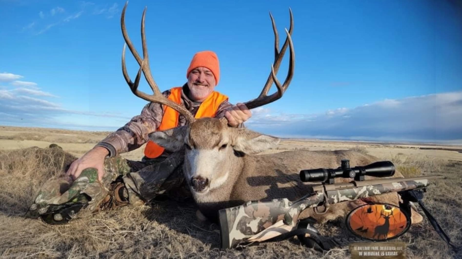 A man in orange vest and hat smiles, holding large antlers above a deer. Rifle with scope lies beside it on field. Clear sky.