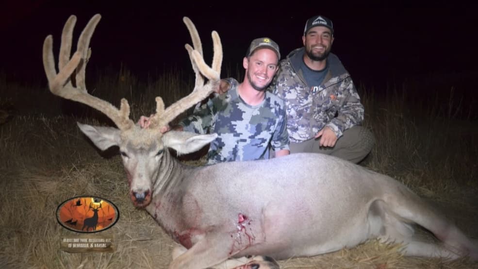 Two men kneel with a large buck with huge antlers. Buck is lying on the ground, wounded with visible blood. Men wear hunt...