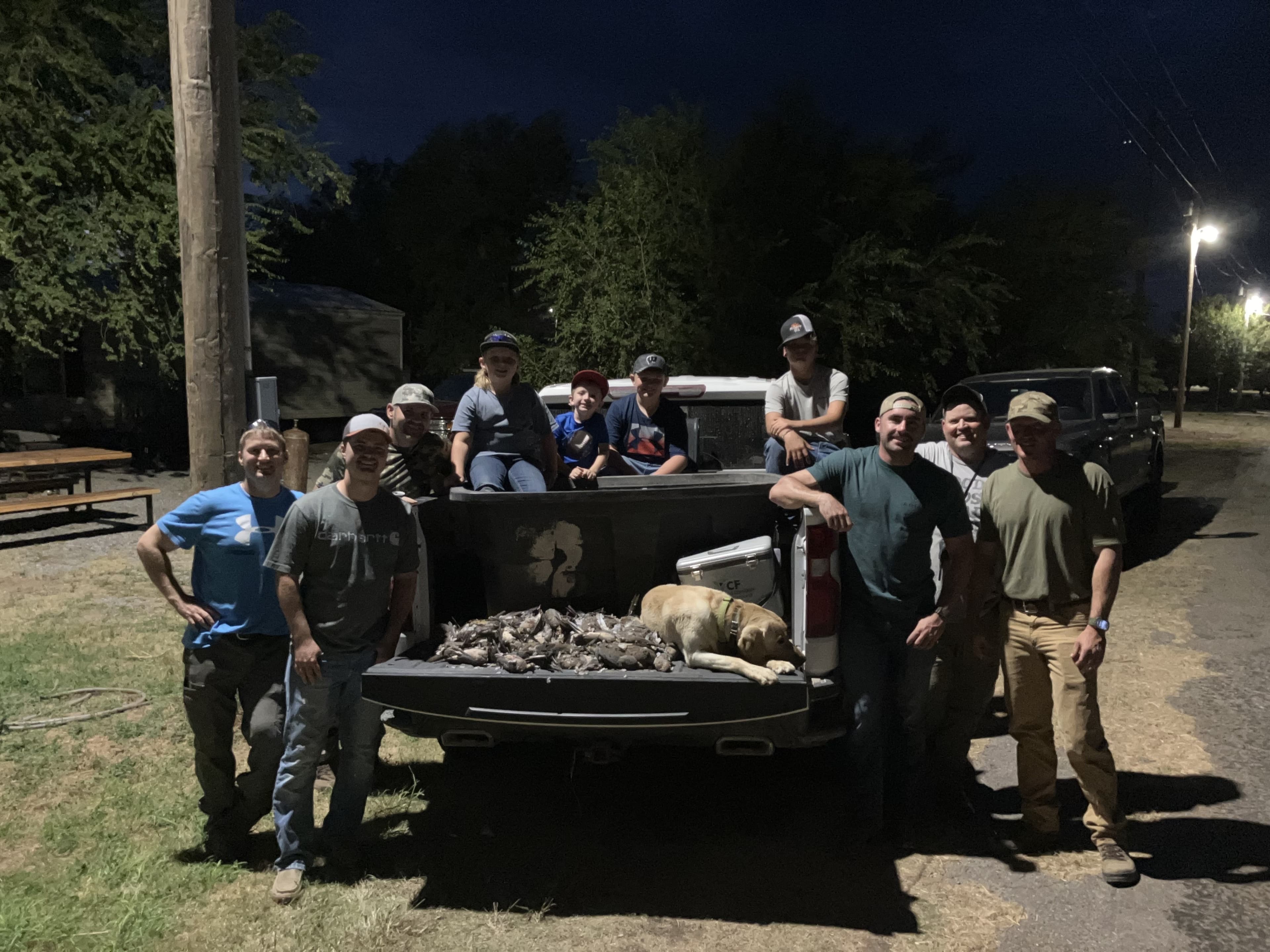 A group poses at night. Men and children stand near a truck bed filled with hunted birds, a dog, and coolers.
