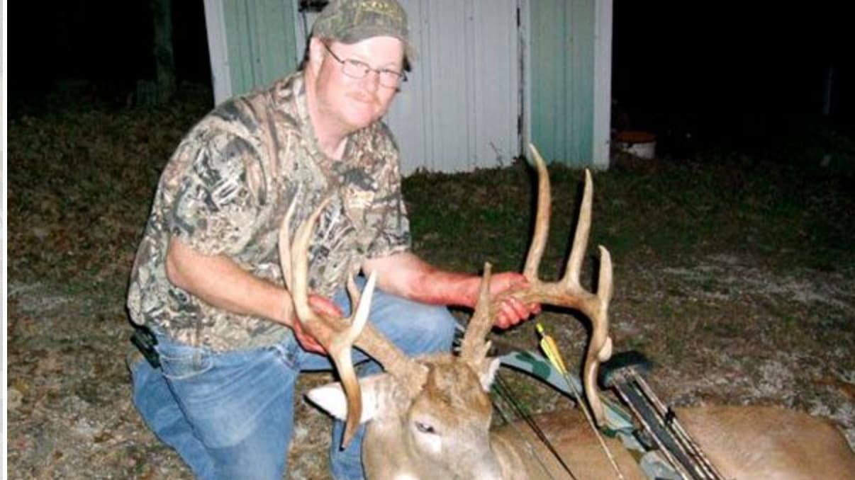 Hunter kneels with large antlered buck, holding antlers. Bow and arrow visible.