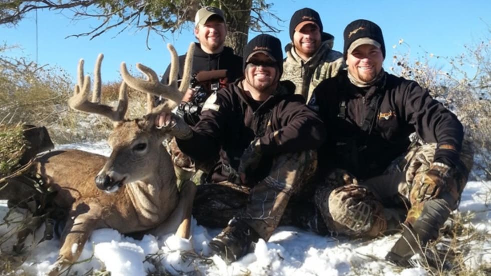 Group of men pose with a large buck in the snow. Deer lies in front of them with large antlers. Men wear hats and camoufla...