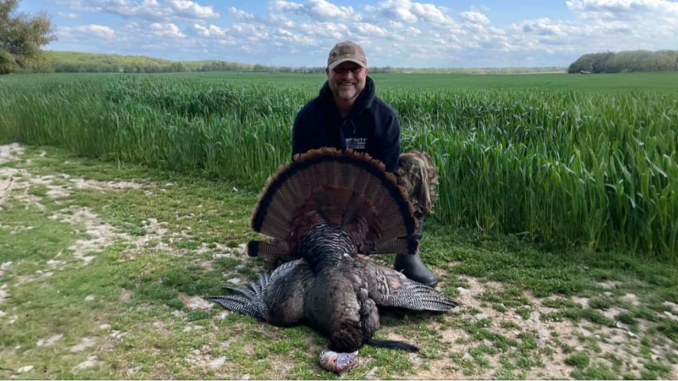 Man kneeling next to a dead turkey on grassy ground, its feathers spread. Field and sky in the background. Man wearing hat...