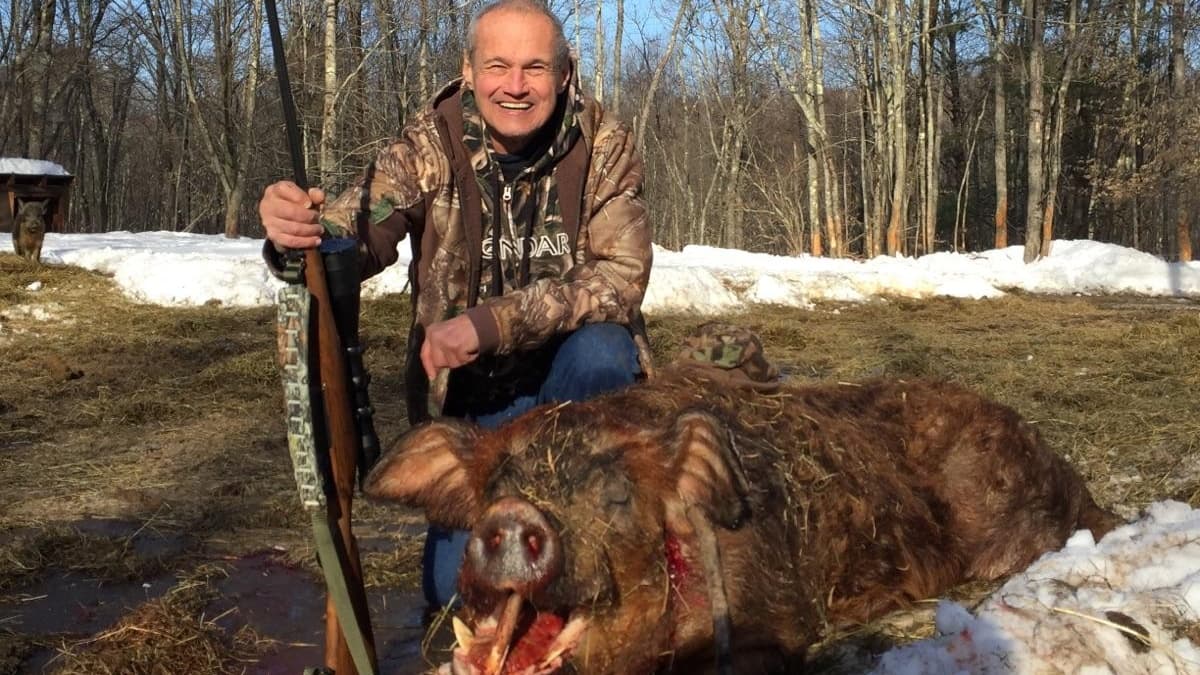Man kneels next to a dead boar, smiling. He holds a rifle; the boar has visible tusks. Background includes snow and trees.