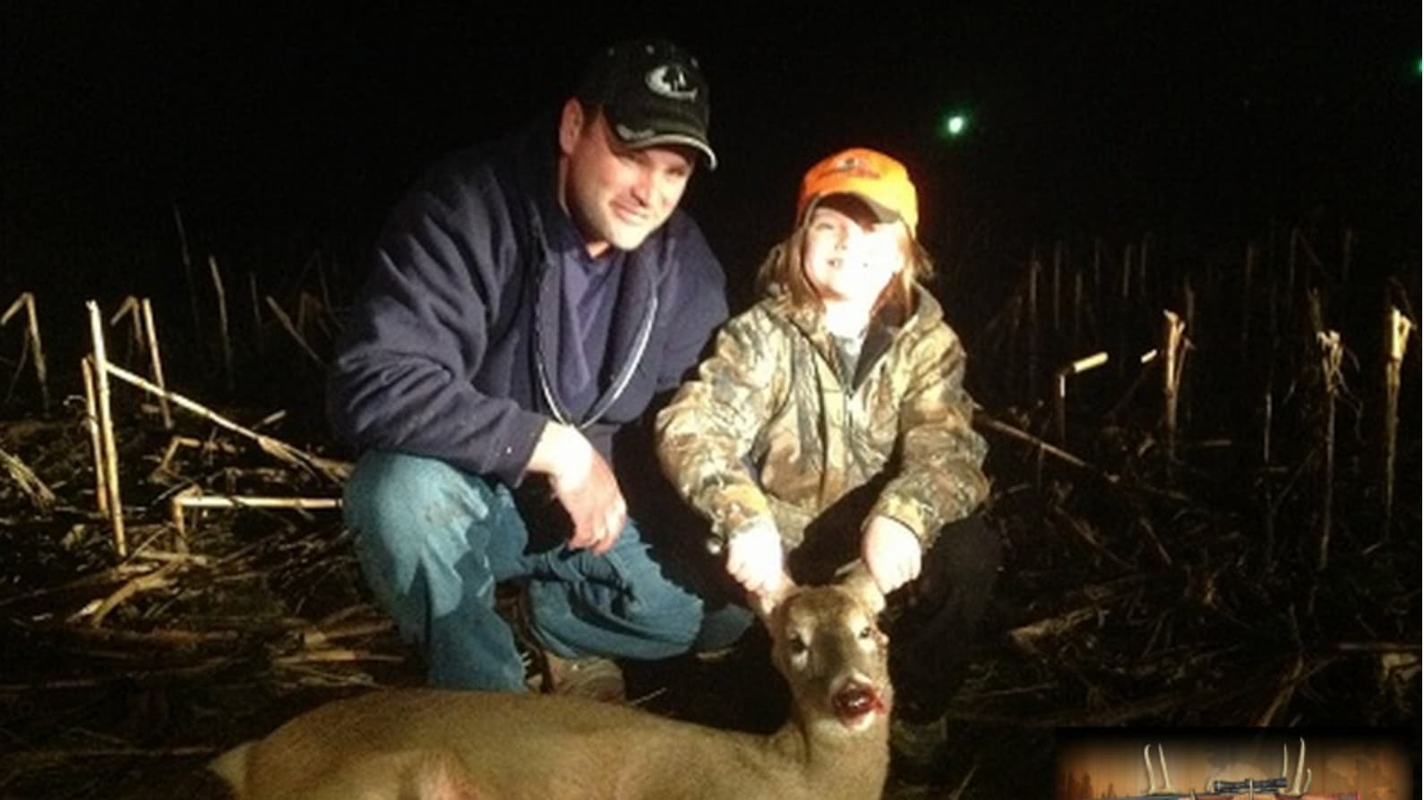Man and child with harvested deer at night in a field with corn stalks.