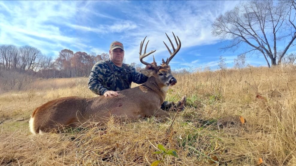 Man in camo, kneels beside large buck with impressive antlers on grassy field. Blue sky with clouds in the background. Aut...