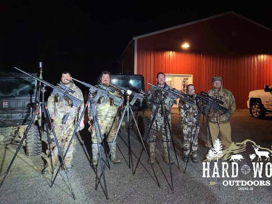 Five people in camouflage stand with rifles on tripods outside a red barn.