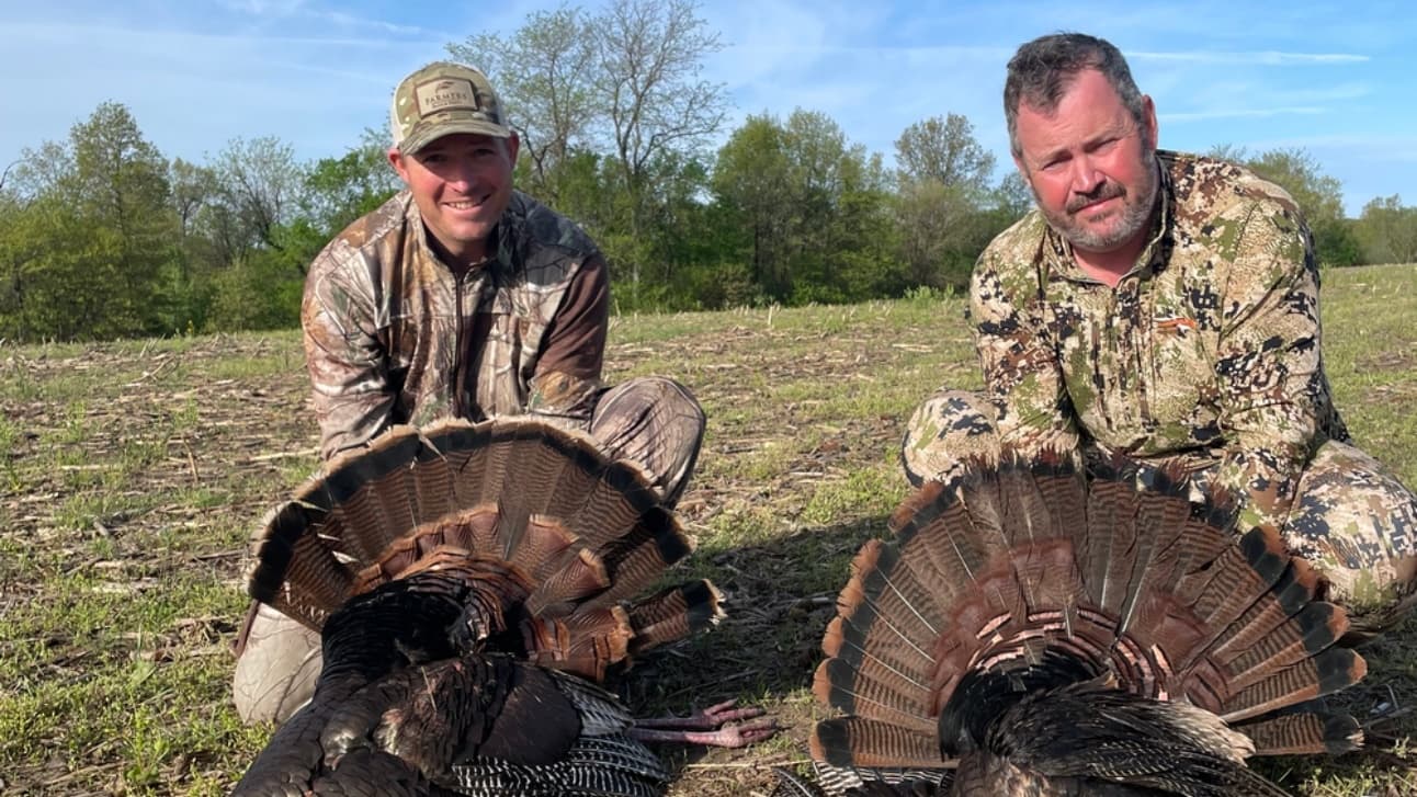 Two hunters in camouflage pose with two large turkeys, their tail feathers fanned out.