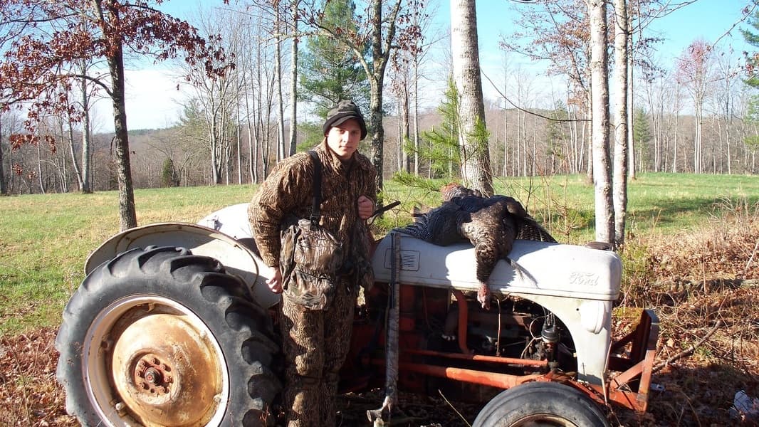 Hunter in camouflage with a harvested turkey by an old Ford tractor in a field.