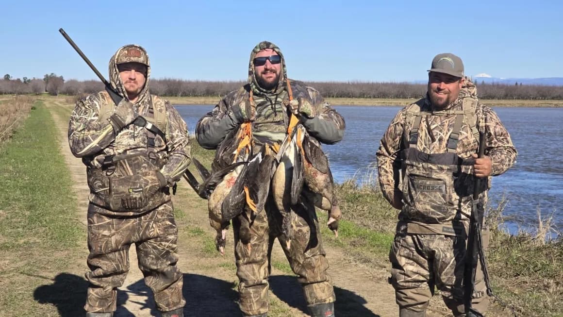 Three men in camouflage hunting gear stand on a path. One holds many ducks. Guns visible. Background is a river and sky.