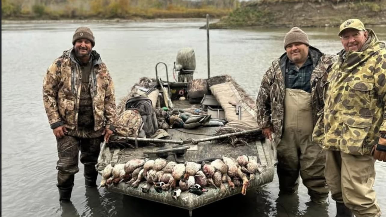 Three men in hunting gear stand near a boat with dead ducks. Boat in water has camo detailing and is loaded with gear.