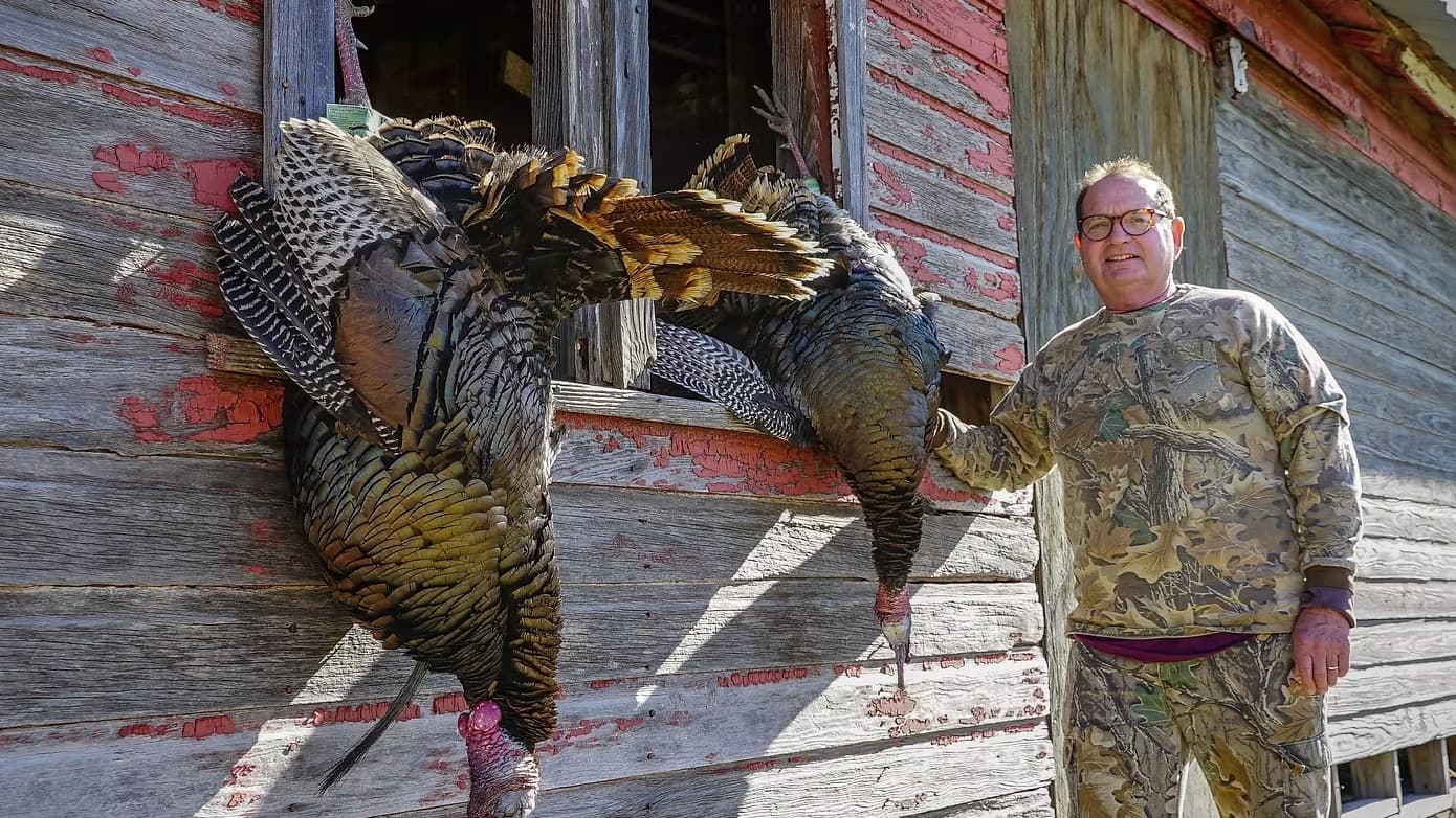 Two wild turkeys hang from an old wooden structure. Man in camouflage next to them, smiling. Buildings have peeling paint.