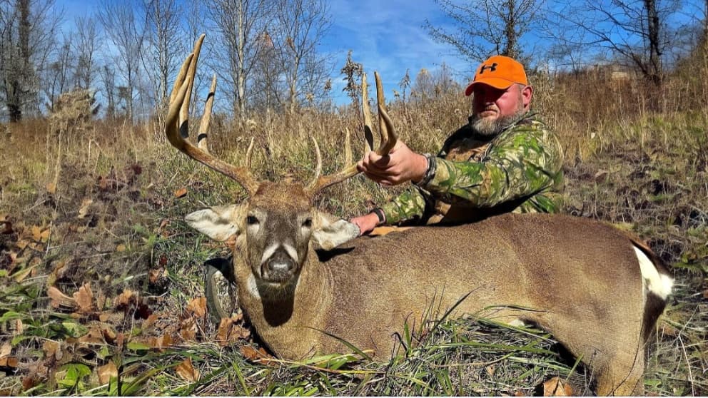 Man in camo holding massive buck's antlers. The deer lays on the ground in a field. Man wears an orange hat. Blue sky and ...