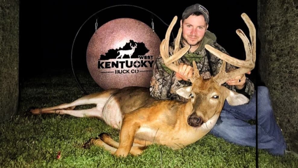 Man with a camouflage jacket and a deer lying on grass. Man holds large antlers; a "West Kentucky Buck Co." sign in backgr...