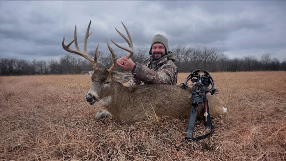 A man in camo and hat smiles next to a large buck with antlers in a field. A crossbow rests nearby. Cloudy sky background.