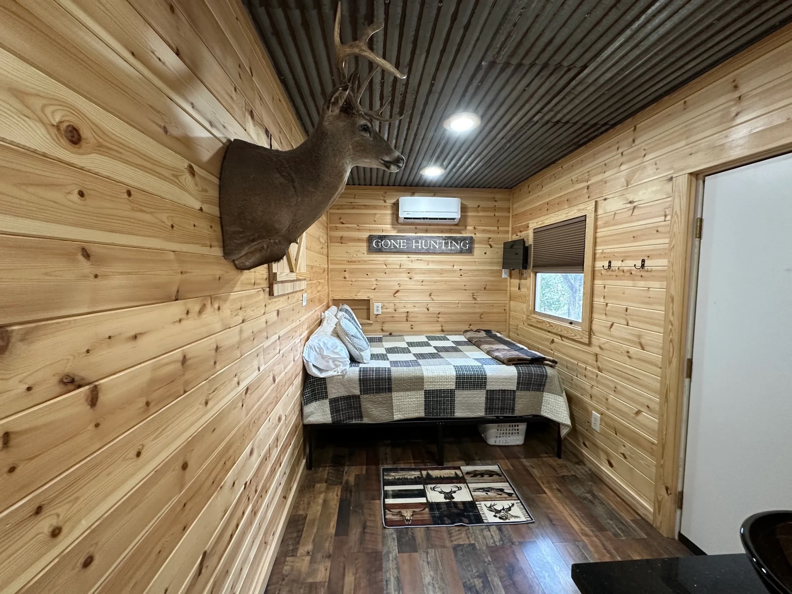 Rustic cabin interior with a deer head mount, bed, and "Gone Hunting" sign. Wood paneling, metal ceiling, and a patterned ...
