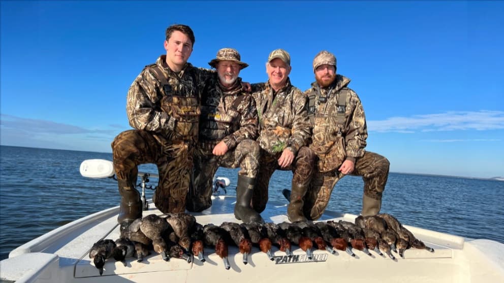 Four men in camouflage gear pose on a boat with a row of ducks. Clear blue sky. Ducks are lined up on the boat's edge.