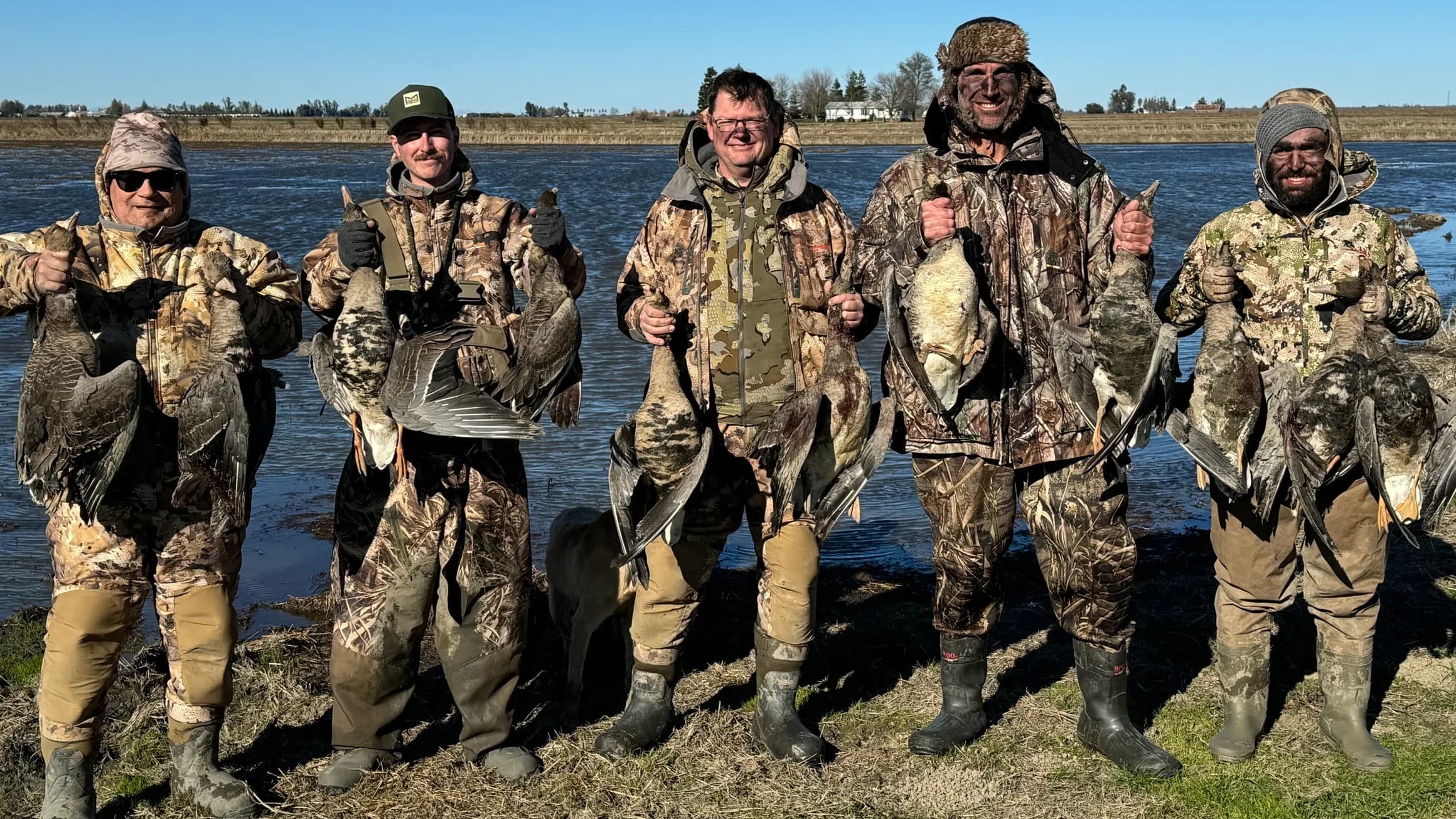 Five men in camouflage, holding ducks, pose by water. All wear waders; three have face paint, and one has a furry hat.
