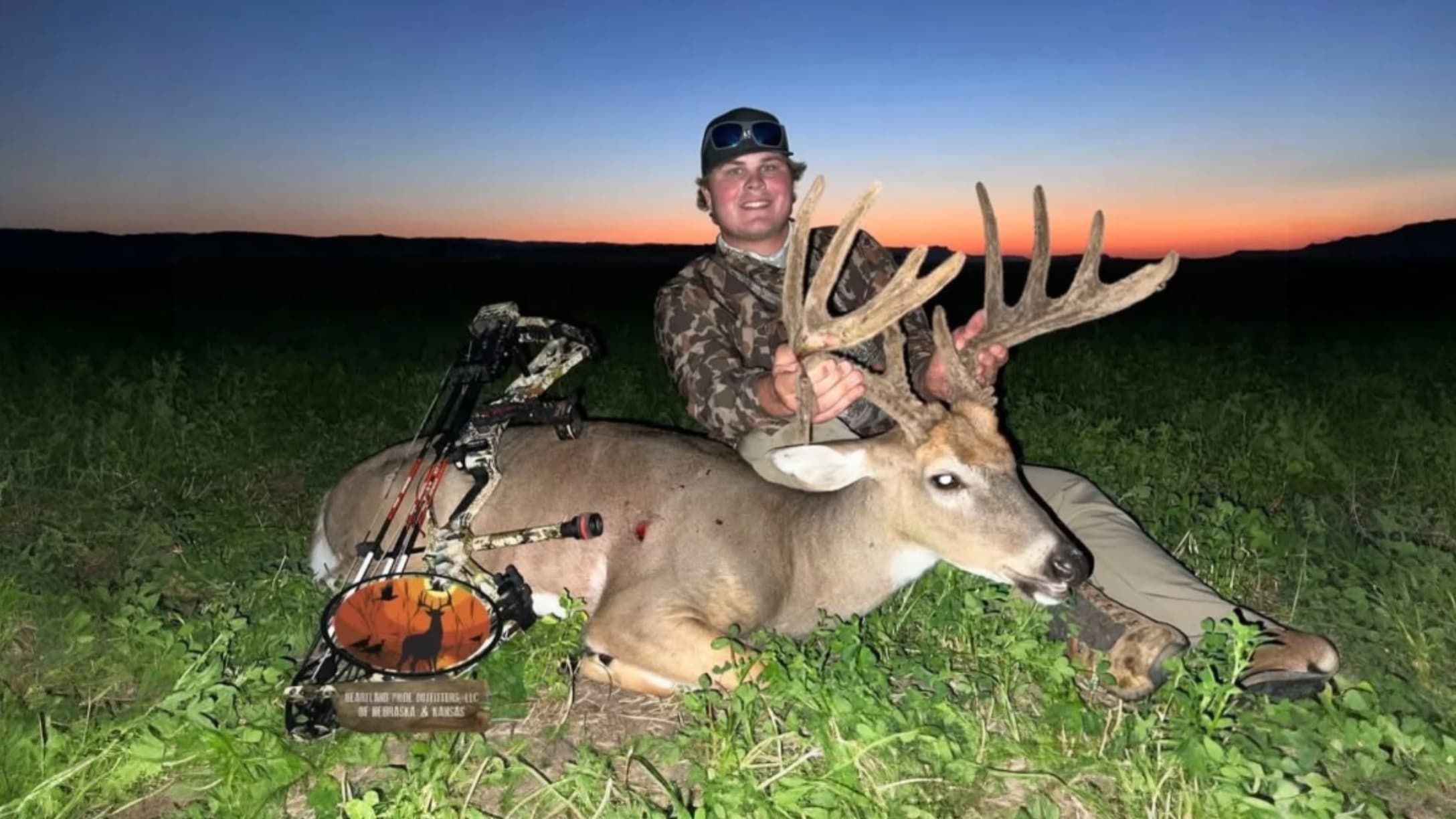 Man with a large antler buck he shot at dusk. He is sitting next to the deer, holding the antlers. A bow rests beside the ...