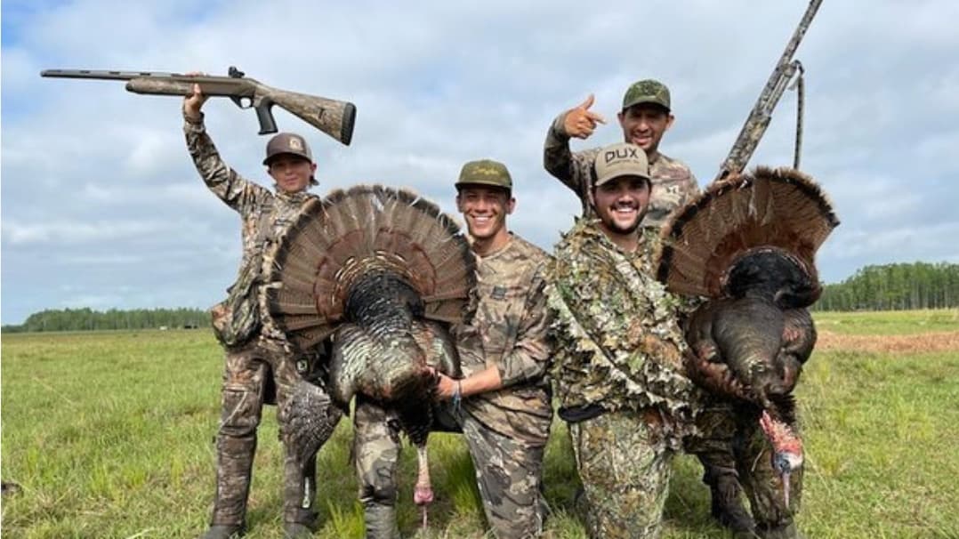 Four men in camouflage with two turkeys they have hunted, holding them up. One man holds a shotgun over his head.