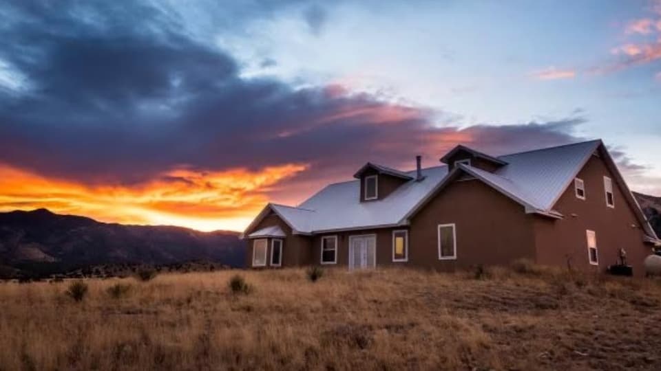 Brown house with a silver roof against a vibrant sunset sky. The house is on a grassy hill, with mountains in the distance.