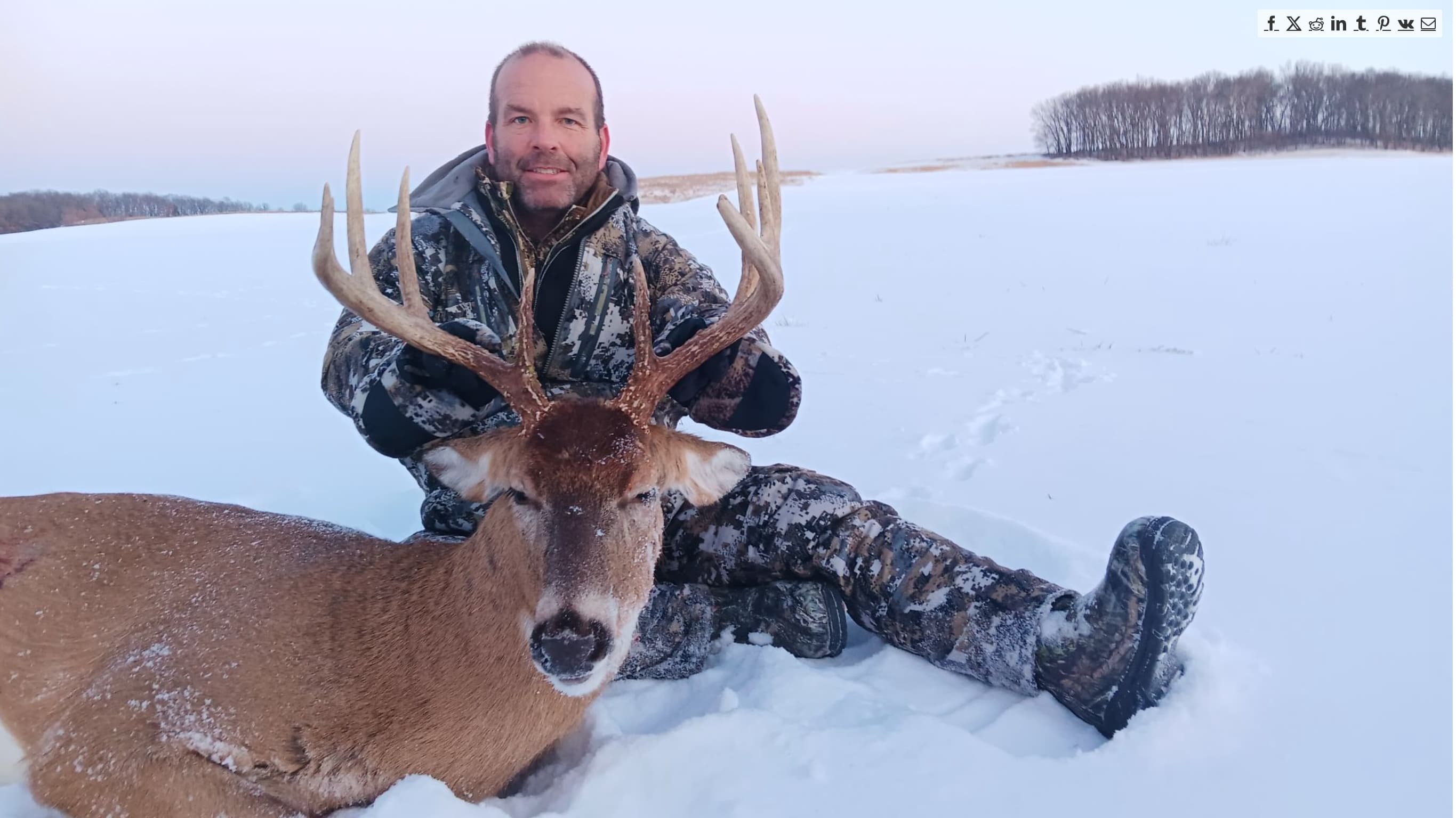 Hunter in camouflage sits next to a buck with large antlers in a snowy field.