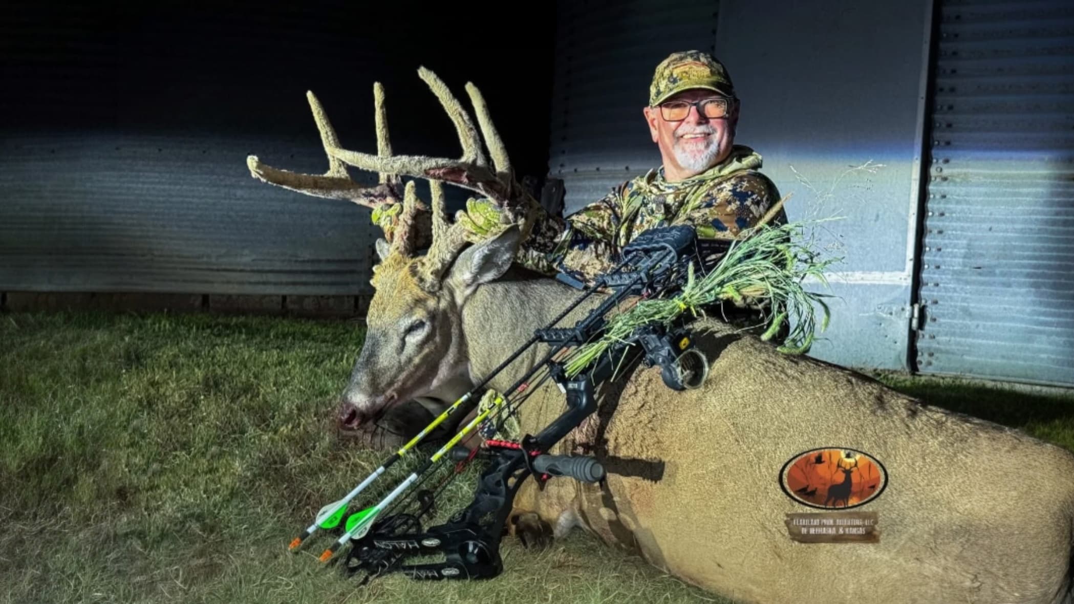 Man in camo with a large buck. Buck has large antlers and an arrow. A bow lies on the deer. Man is smiling and holding veg...