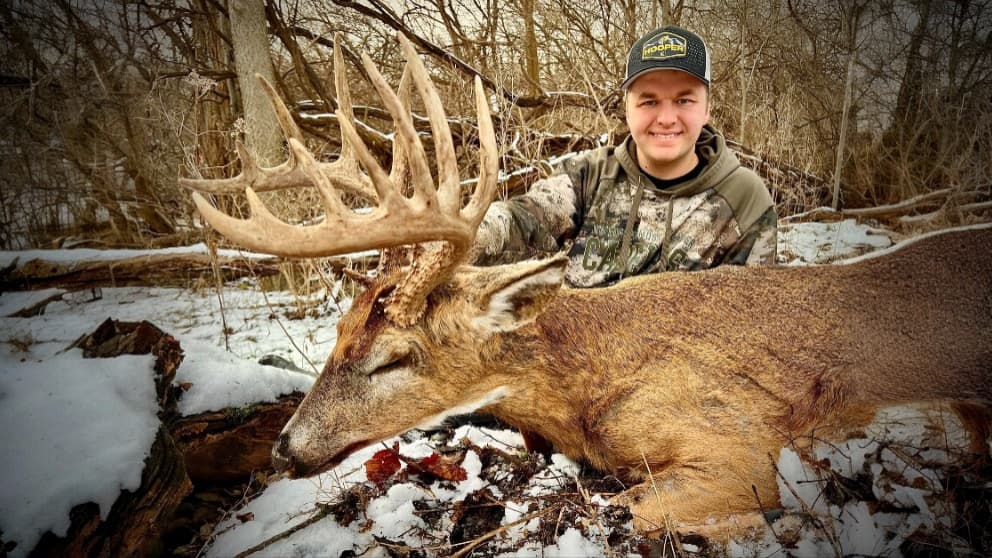 Smiling man in camo and hat with a large buck. The buck has a massive rack of antlers and lies in snow. Bare trees in back...