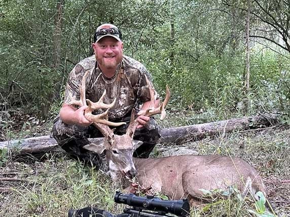 Hunter proudly displays a large antlered buck in a wooded area.
