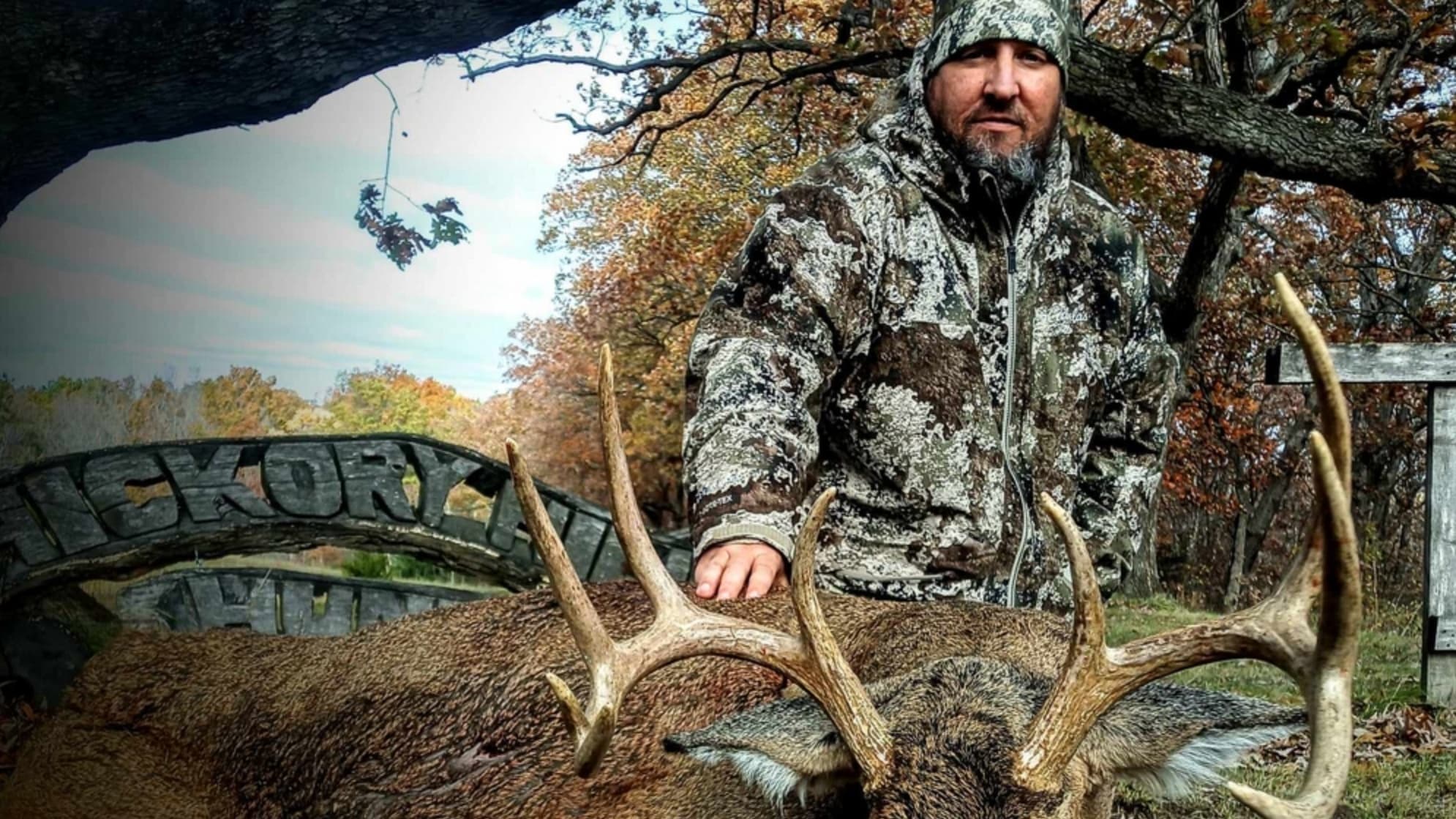 Hunter in camo jacket posing with a large antlered deer. Wooden "HICKORY" sign in background.