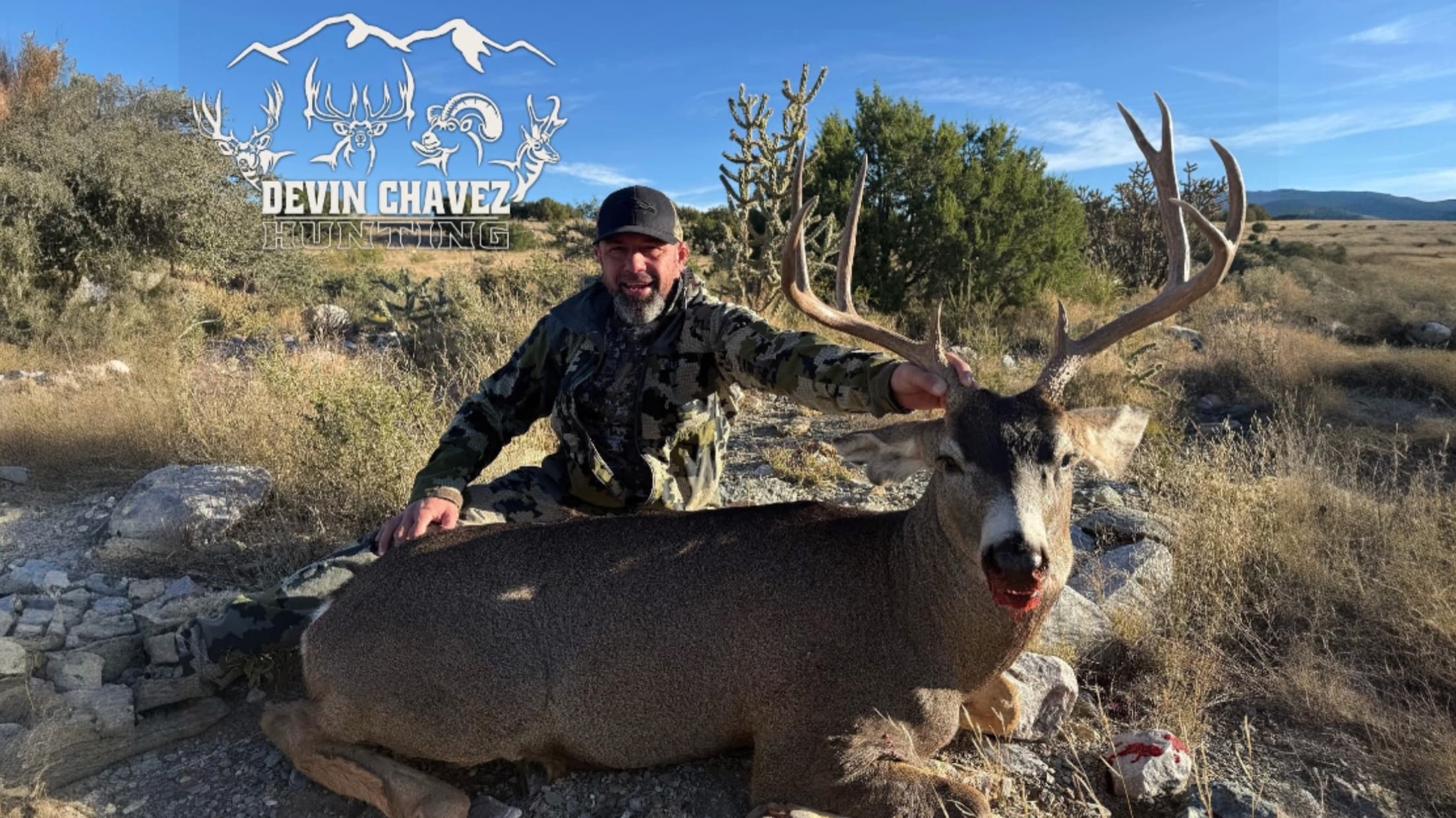 Man in camo kneels beside large buck with impressive antlers. Buck is lying down, blood on its face. Background is dry gr...