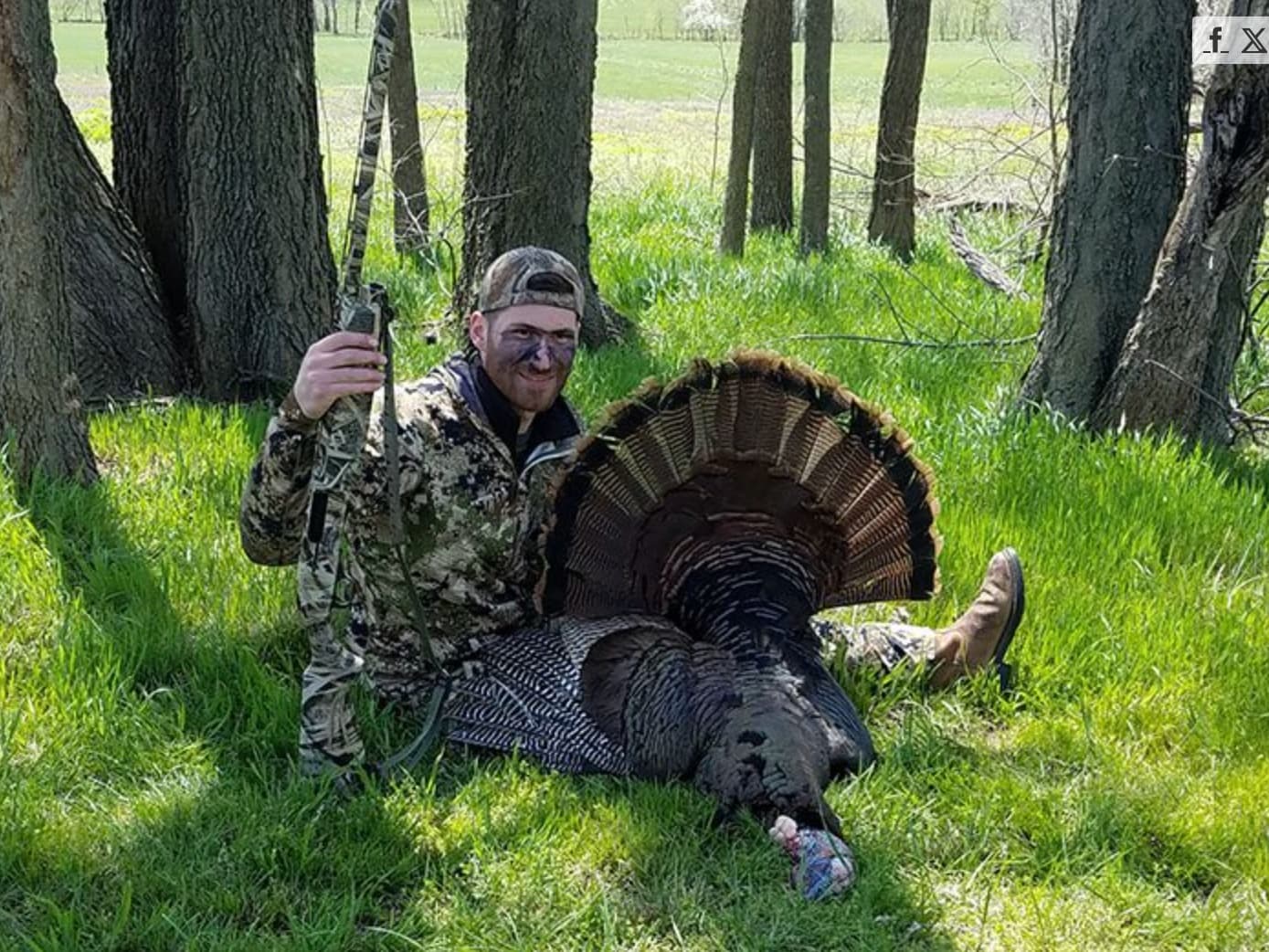 Hunter in camouflage with face paint smiles and holds shotgun next to a large harvested turkey with fanned tail feathers.