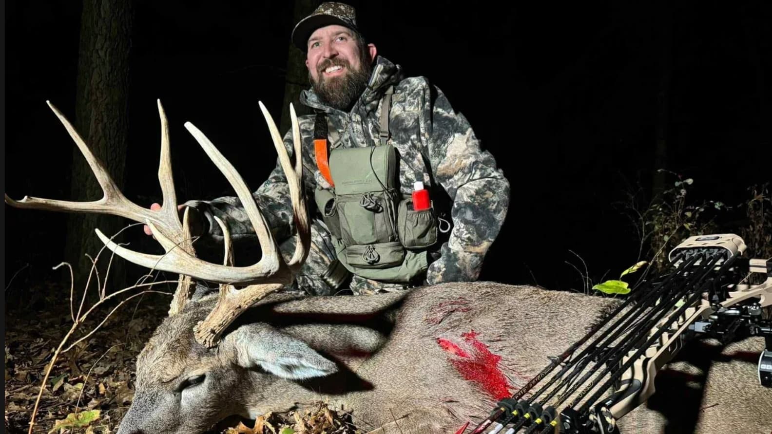 Hunter smiles with a large buck. Buck lies on forest floor; blood on side. Bow in frame. Hunter wears camo, orange strap.