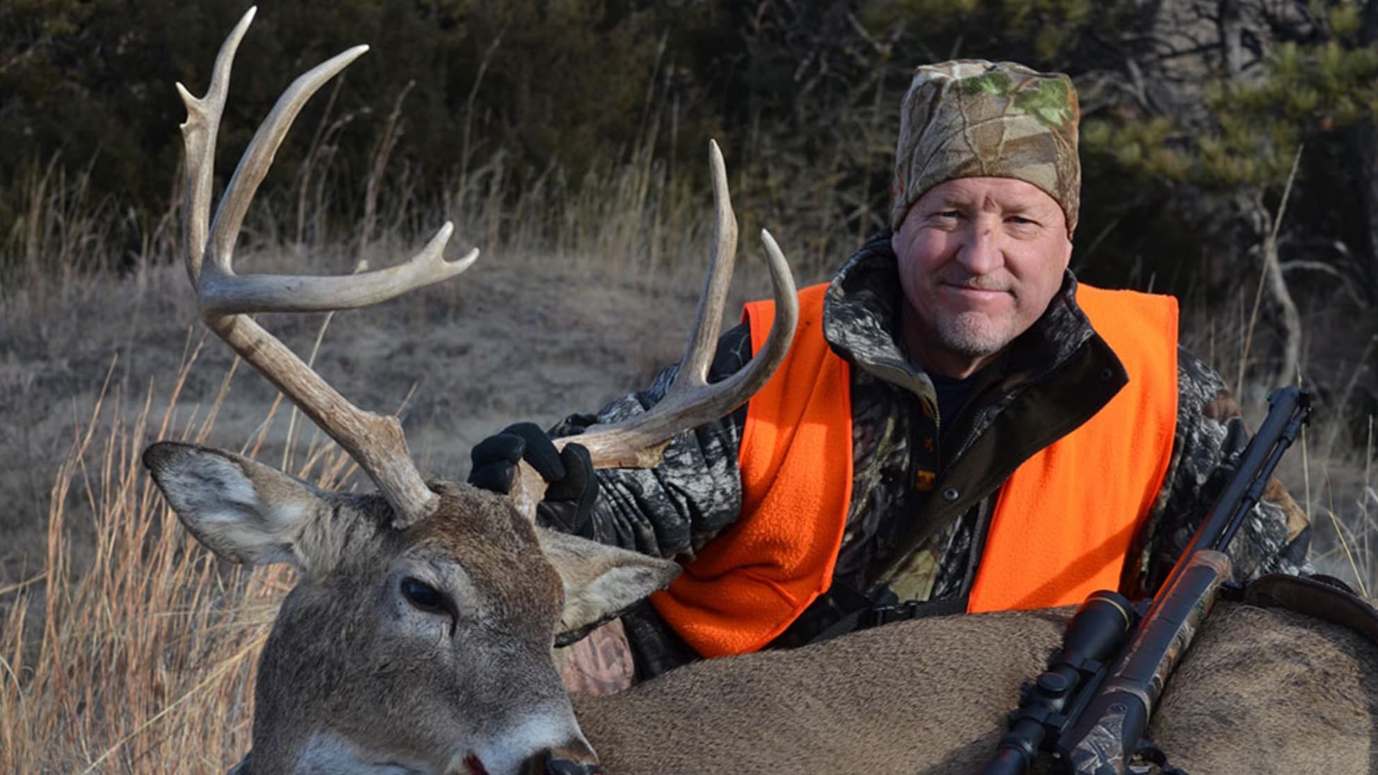 Man in camouflage, orange vest, posing with a harvested deer. Deer has large antlers, resting next to rifle. Smiling at c...
