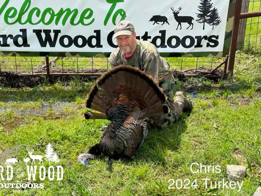 Man kneels, holding a wild turkey with fanned tail feathers, in grassy area. Welcome sign behind. "Chris 2024 Turkey" text...