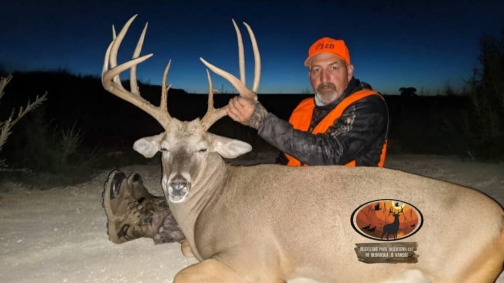Hunter with a large buck. Hunter in orange vest and hat holds buck's antlers. Buck lies on ground. Logo present on buck.