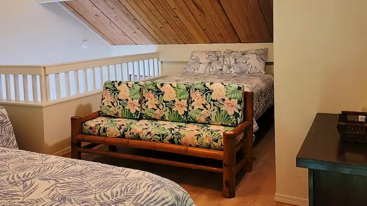 A cozy bedroom with a wooden framed sofa covered in floral cushions and a bed with leaf patterned bedding. Wooden ceiling.