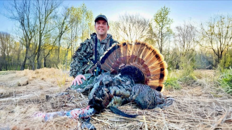 Man in camouflage with a harvested turkey. The bird's fanned feathers are visible. The scene is outdoors with trees in the...