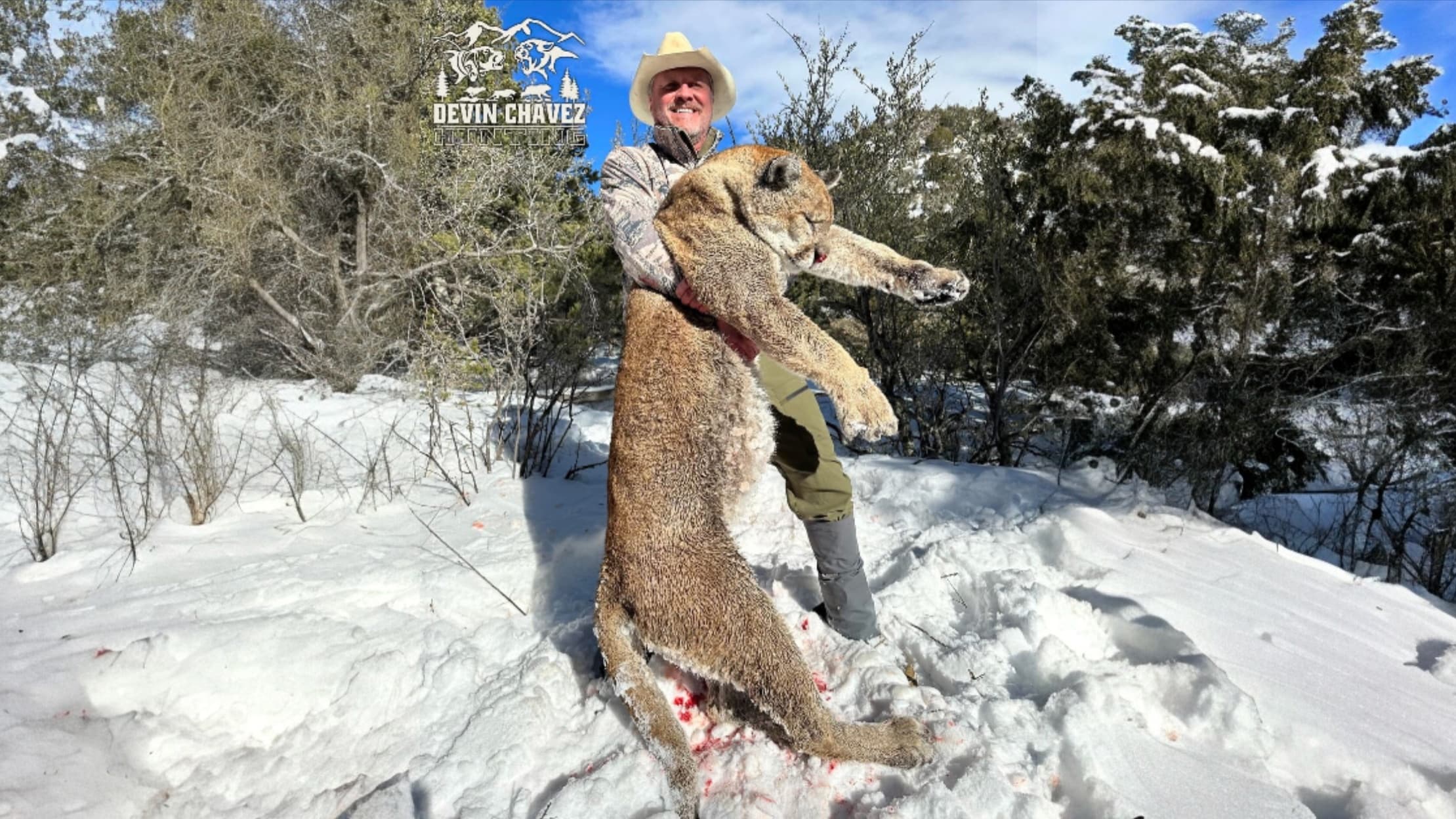 Man in cowboy hat holding a large dead cougar in snowy woods. The cougar’s body is covered in blood. Background: trees and...