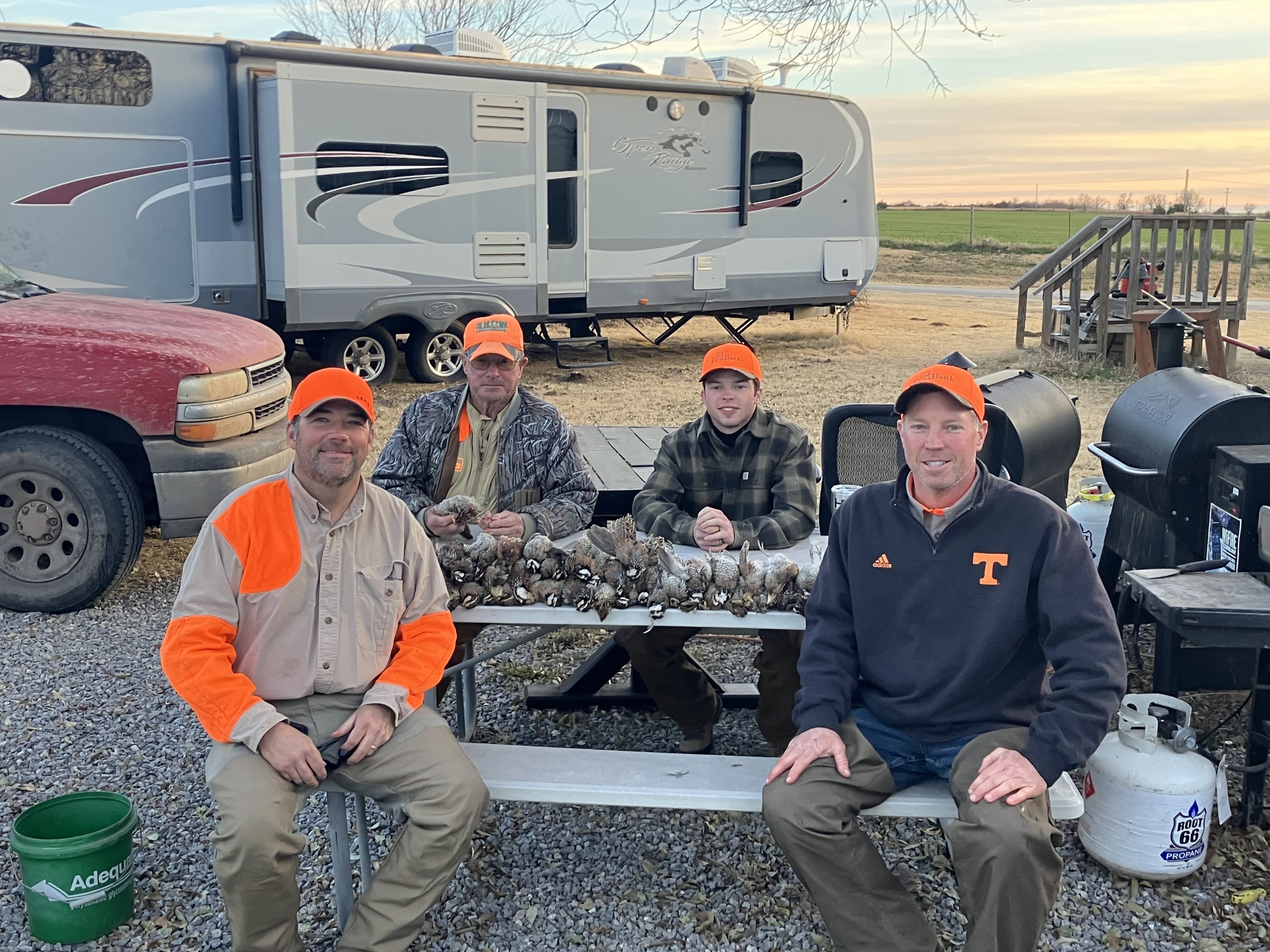 Four men in hunting gear sit by a table with harvested birds. A trailer and red pickup truck are in background. A gas gril...