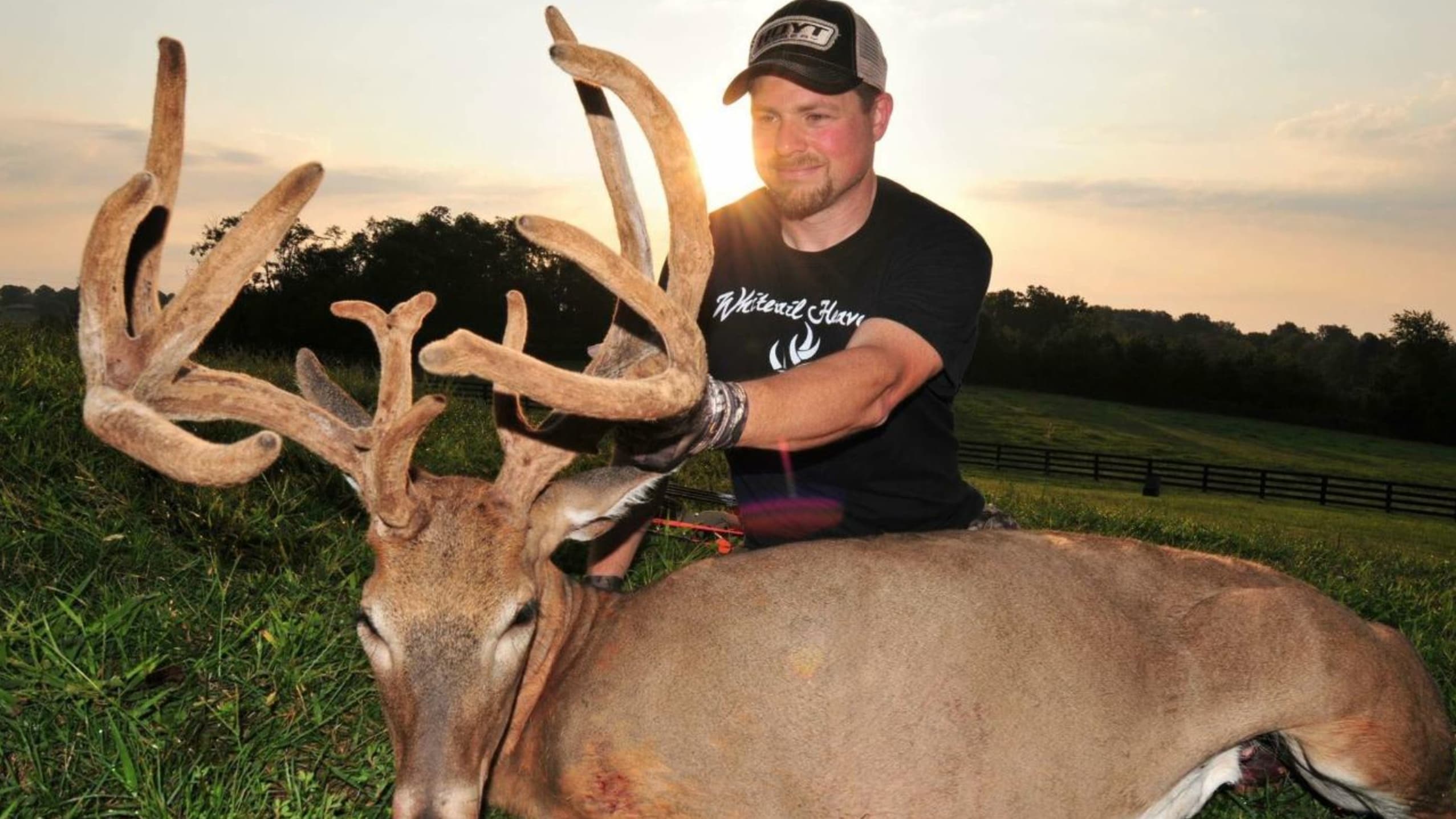 A man kneels beside a large buck with impressive antlers. The deer lies in green grass. The setting sun casts a warm glow.