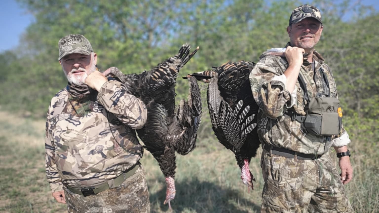 Two men in camouflage carry harvested turkeys over their shoulders. One turkey per man. Both men are smiling, outdoors.