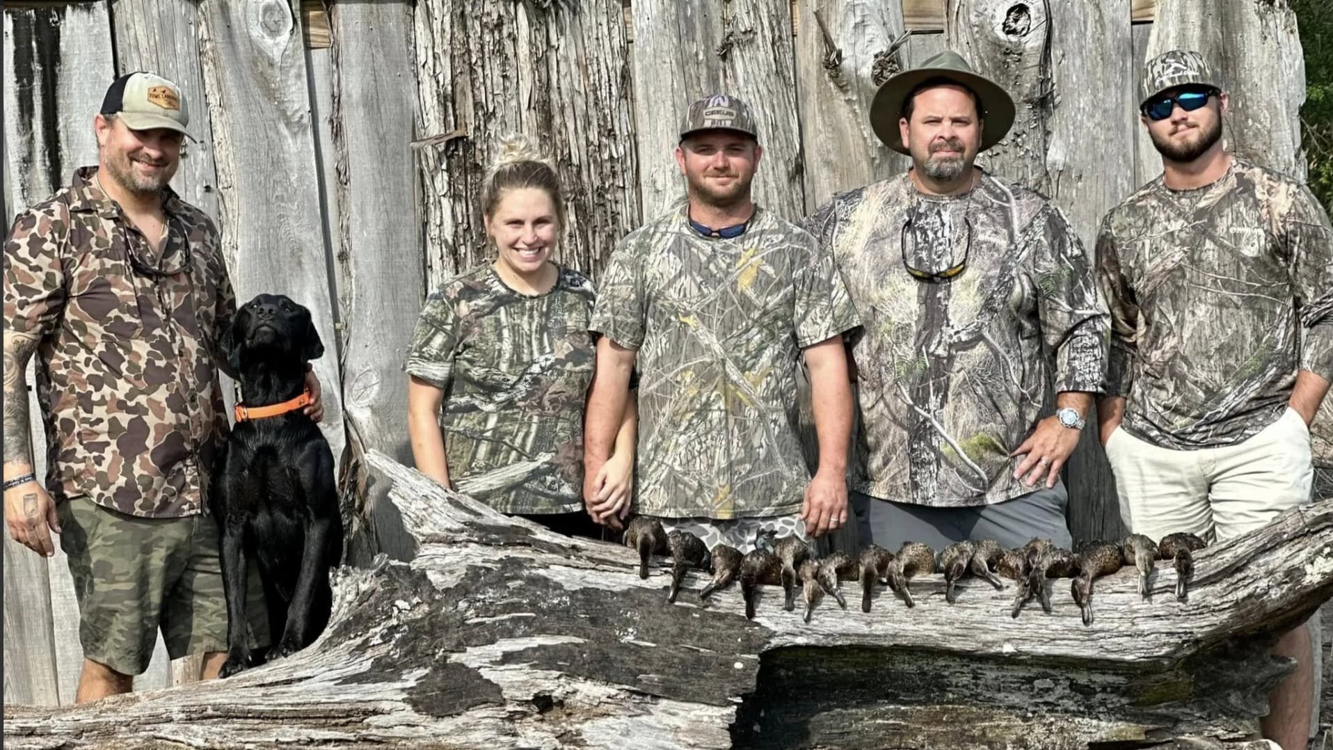 Five people in camouflage clothing pose with a black dog and harvested ducks on a log in front of a wood wall.