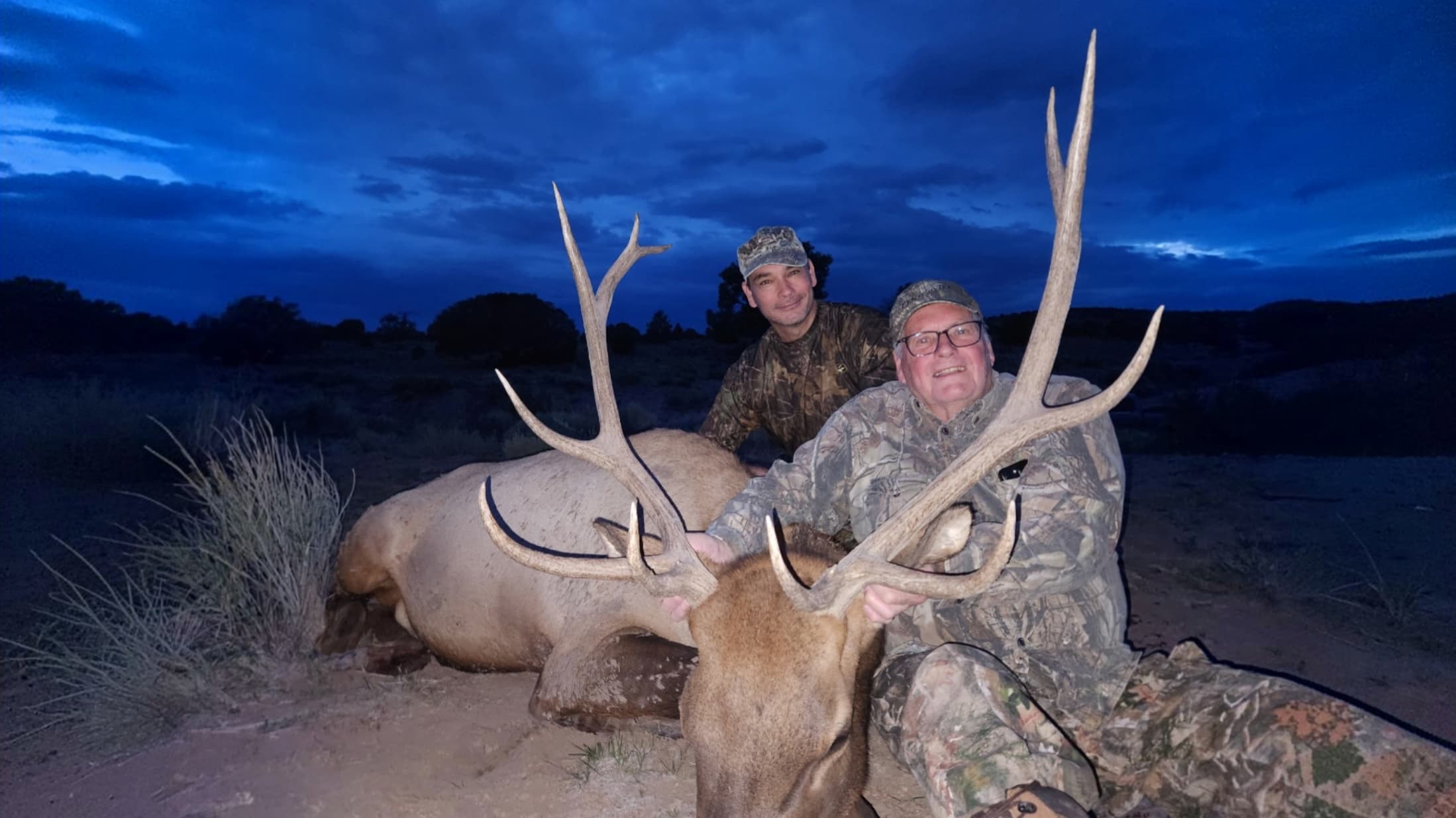 Two hunters in camouflage gear pose behind a large bull elk at dusk.