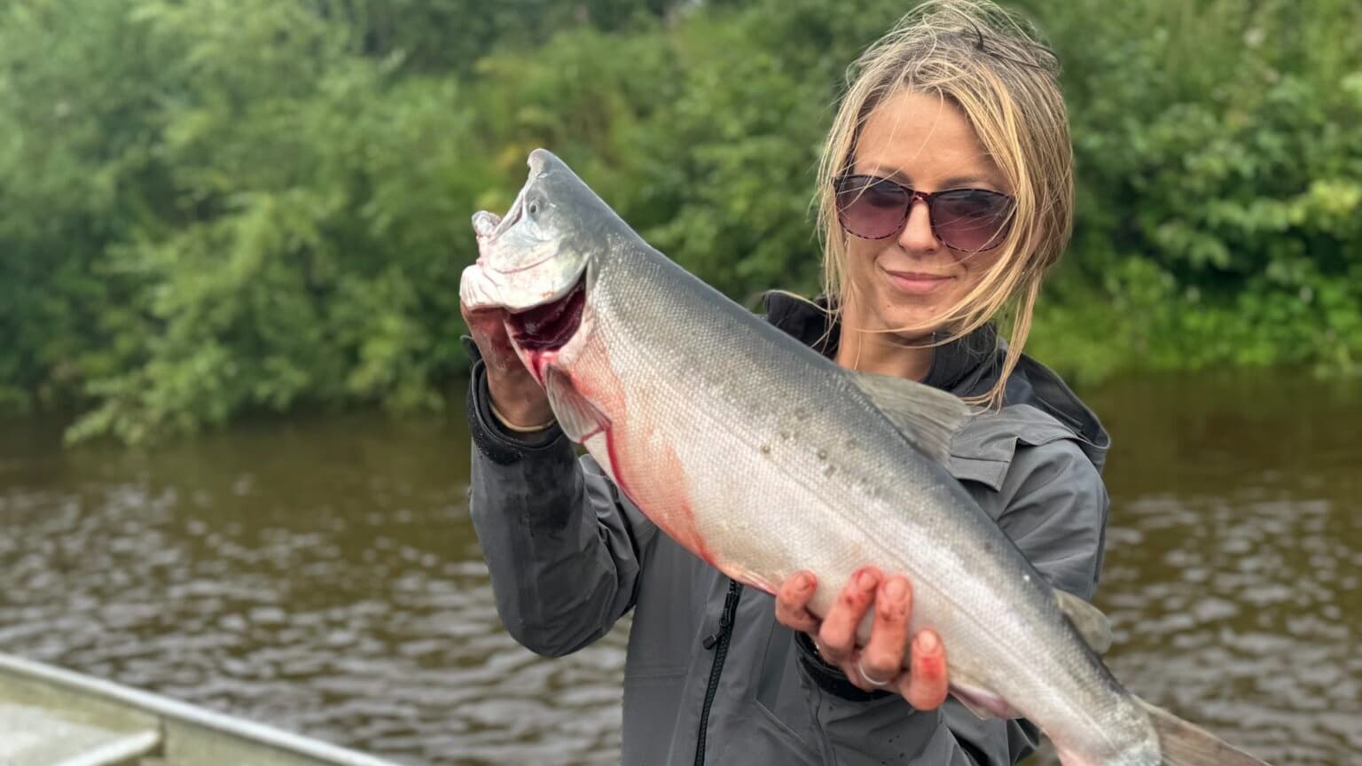 Woman holding a large fish with red gills, outdoors near water. She wears sunglasses, a grey jacket, and smiles. The fish ...