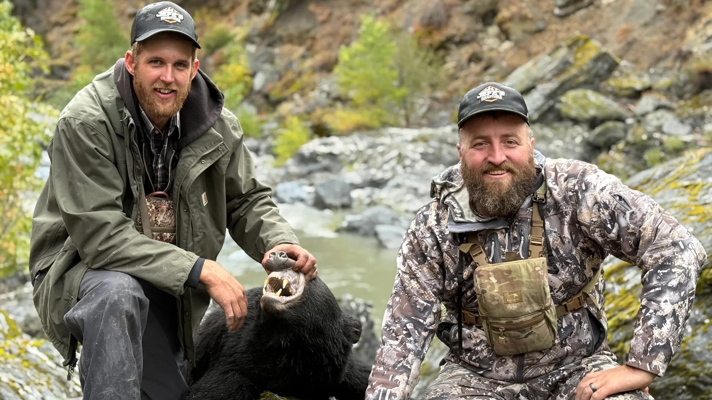 Two men with beards pose with a black bear, its mouth open. One man in green jacket, the other in camo, both wear hats. Na...