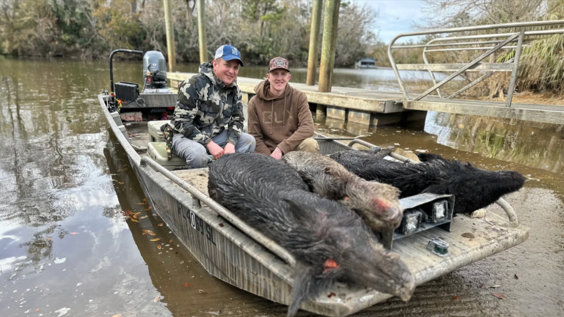 Two men on a boat, posing. Several wild boars are in the boat's bed. The boat sits on muddy bank by a dock and a waterway.