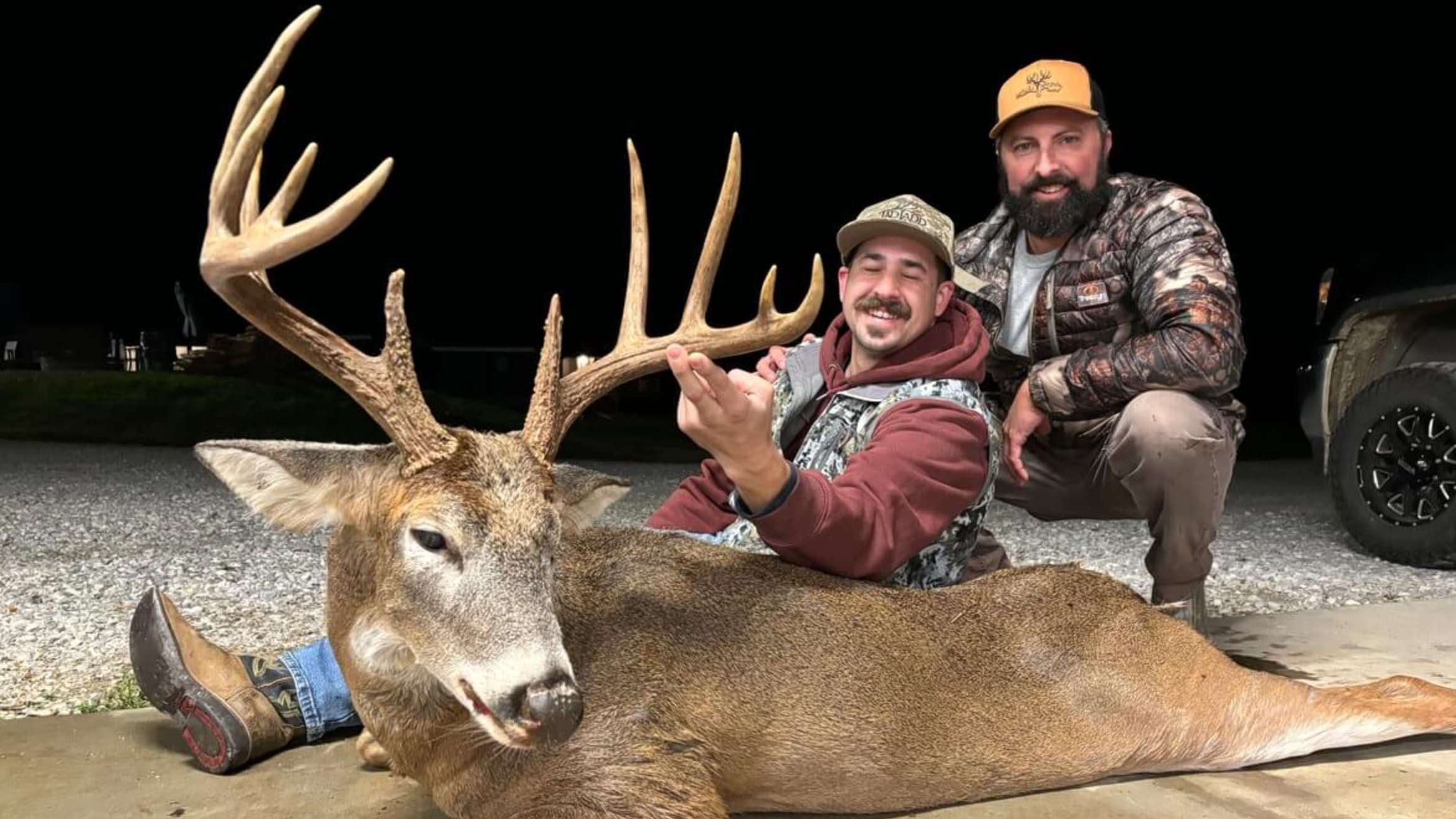 Two men crouch near a large buck with impressive antlers. One man sits on the deer, smiling, while the other kneels, both ...